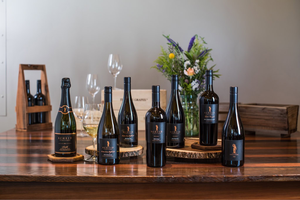 Scheid Vineyard wine bottles with glasses and floral arrangement on wooden table.