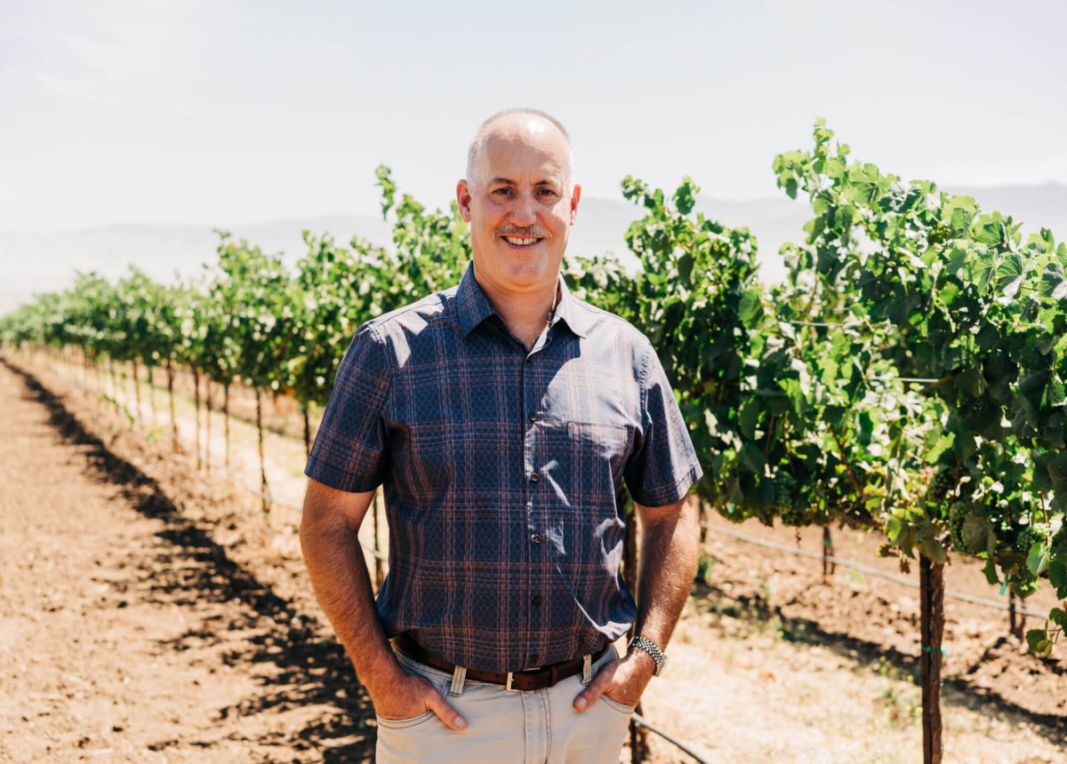 Vineyard owner smiling with lush grapevines at Scheid Vineyards in a scenic outdoor setting.